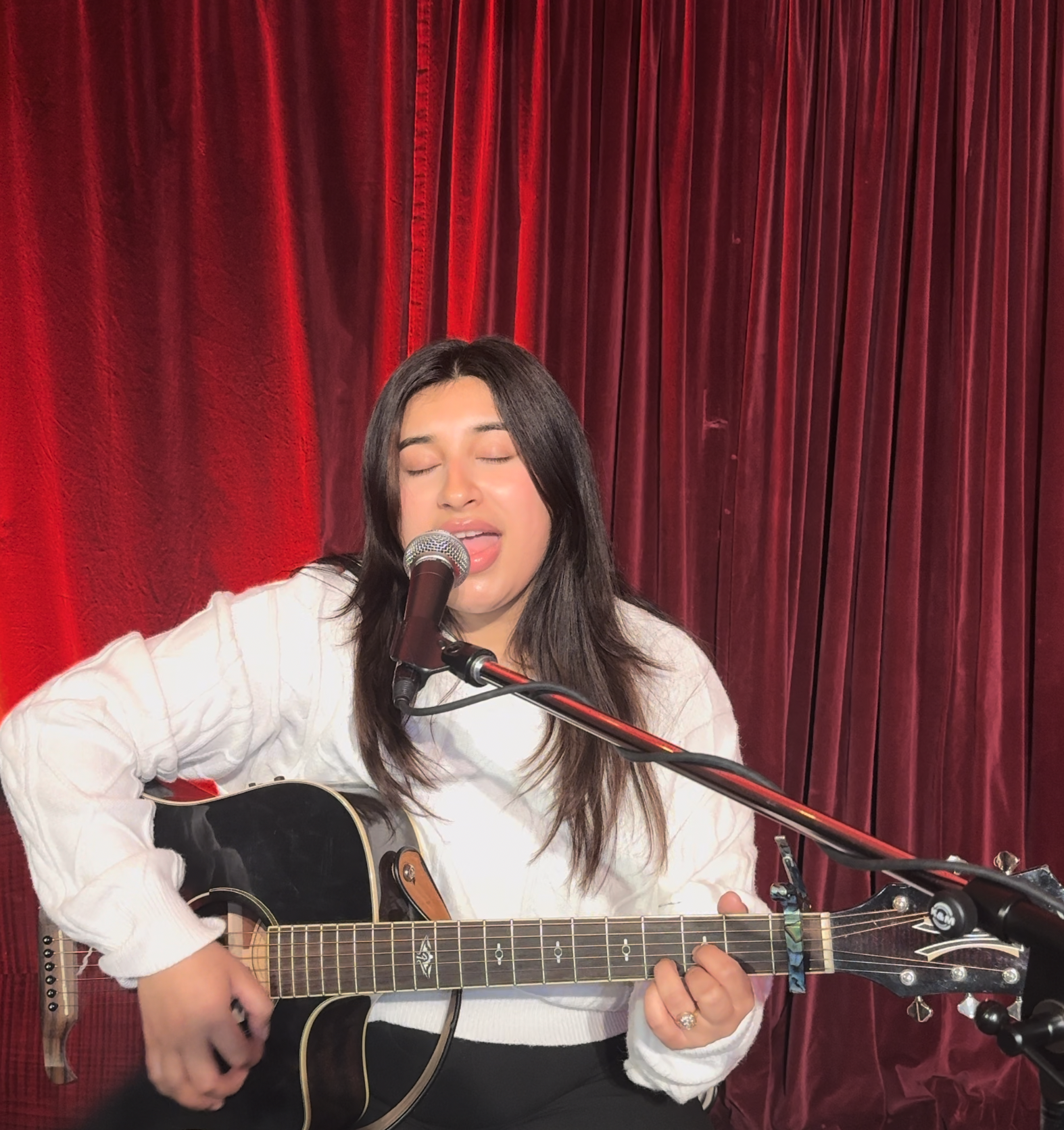 Manvi Thakur singing and playing guitar against a red curtain